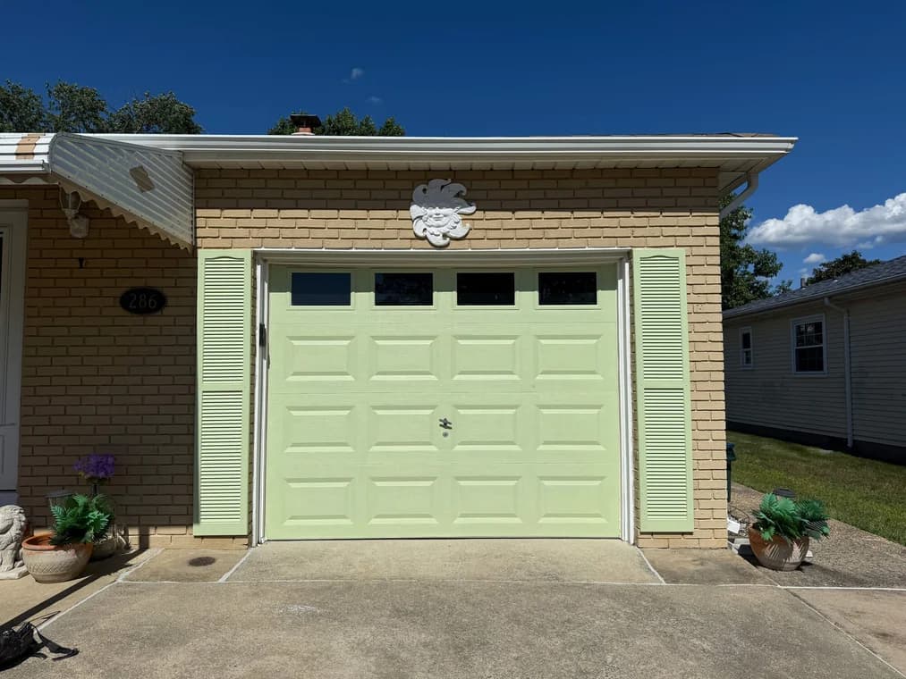 This is an image from Carfagno Professional Painting & Power Washing LLC. It shows a freshly painted light green garage door with matching shutters on a tan brick house.