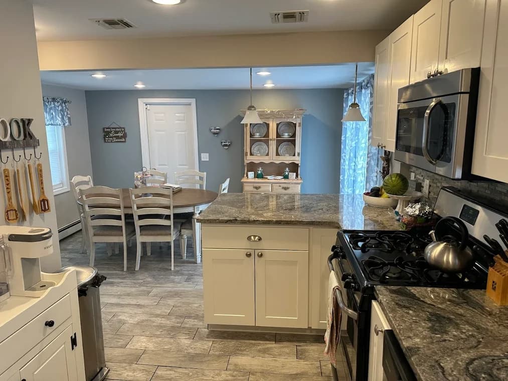 This is an image from Certified Painting, located in New York. The photo shows a kitchen and dining area with light gray walls, white cabinetry, stone countertops, and wood-look tile flooring.
