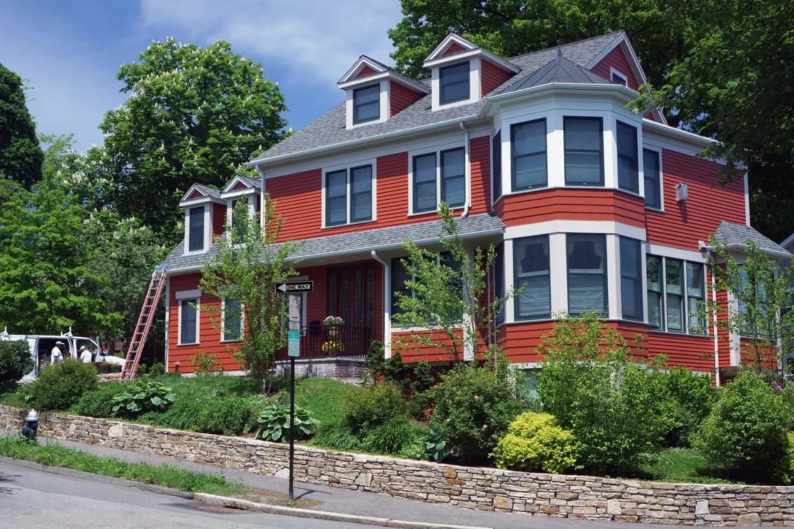 This is an image from Catchlight Painting, located in Massachusetts. It shows a red-painted house with white trim and dormer windows, enhanced by lush landscaping and a stone retaining wall.