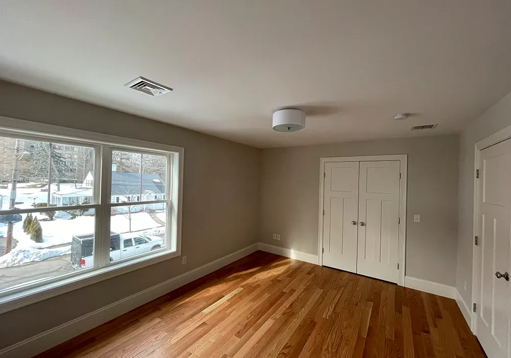 This is an image from Zuck Painting, located in Massachusetts. It displays a freshly painted interior room with soft beige walls, white trim, hardwood floors, and a large window.