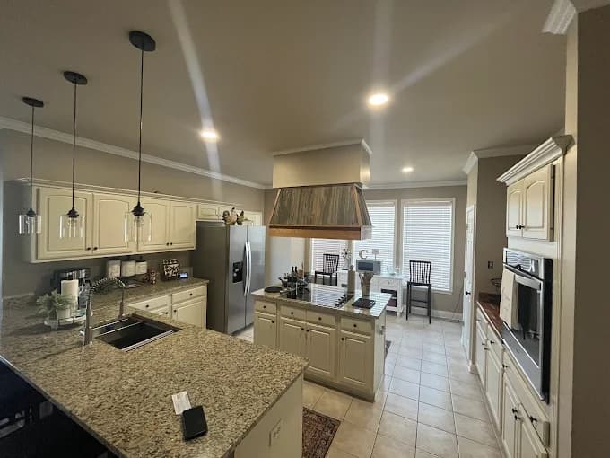 This is an image from Spell Painting, located in Louisiana. The photo shows a renovated kitchen with freshly painted cream cabinets, granite countertops, pendant lighting, and a wood-accented range hood creating a warm, inviting space.