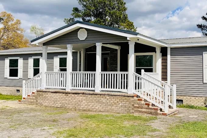 Exterior painting project by Of The Essence Design Build Paint in Wilmington, NC. The image shows a gray home with white porch railings, dark shutters, and brick steps.
