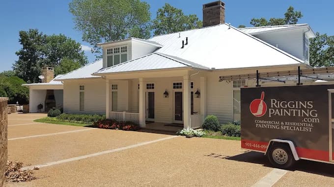 Exterior painting by Riggins Painting in Bartlett, Tennessee. The image shows a white home with a metal roof, covered porch, and branded work trailer.