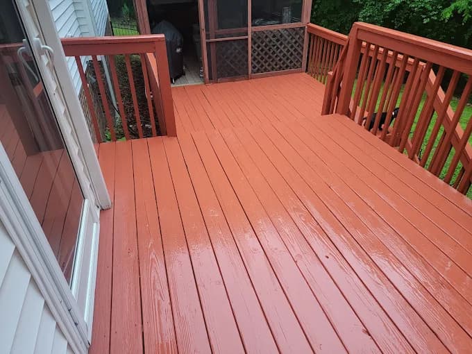 Deck staining project by NC Paint & PowerWash in Garner, North Carolina. The image shows a reddish deck with railings, steps, and screened porch doors.