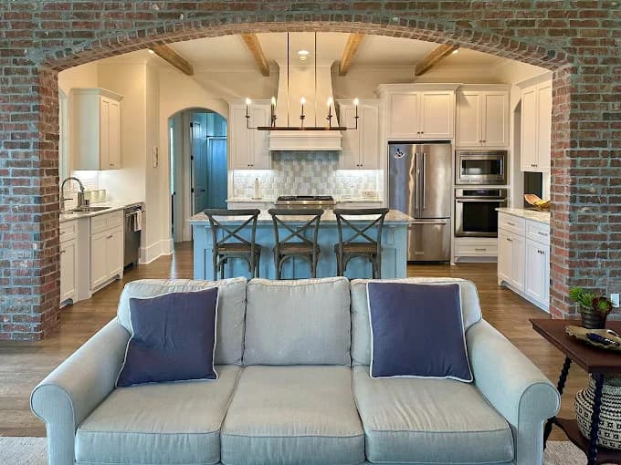 This is an image from 360 Painting of Lafayette, located in Louisiana. The photo shows a beautifully painted open-concept kitchen and living area with white cabinets, exposed wooden ceiling beams, and a brick archway framing a modern light gray sofa.