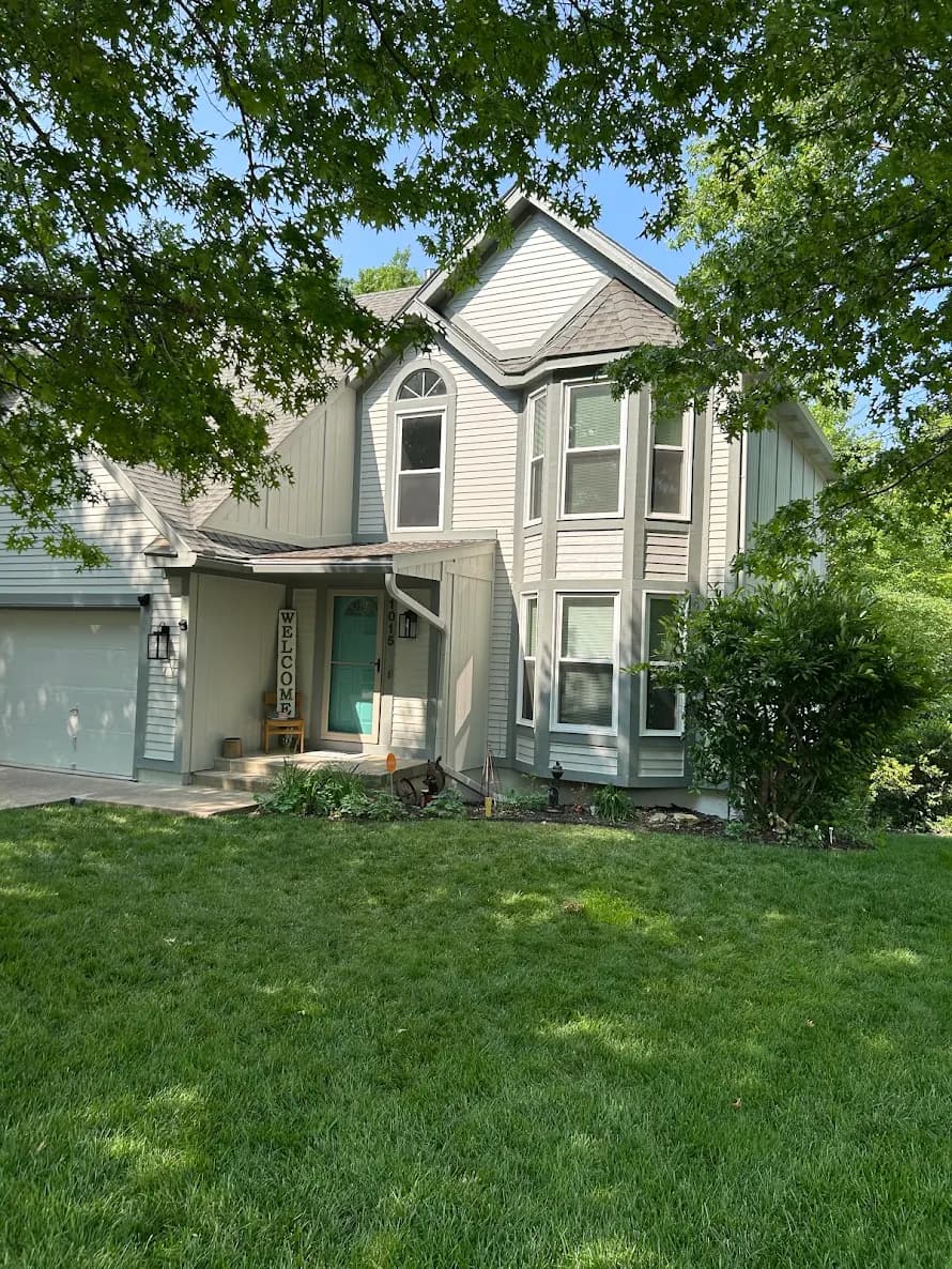 This is an image from Odyssey Painting. The image shows a light gray two-story home with green shutters, a covered entryway, and mature trees providing shade over the front yard.