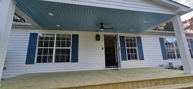 This is an image from Todd's Painting, located in New York. The photo shows a freshly updated front porch with white siding, blue shutters, a light blue beadboard ceiling, recessed lighting, and a new unfinished wood deck.