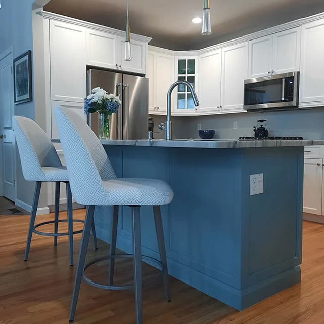This is an image from Linda Loo Designs Cabinet Refinishing & Refacing, showing a kitchen island with light blue cabinetry and white countertops. The room includes two high-backed chairs and white upper cabinets for contrast.