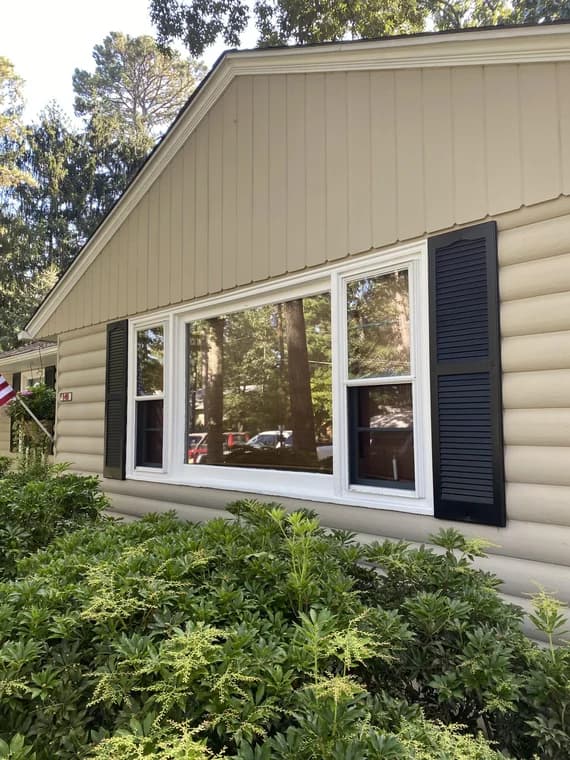This is an image from Pizzazz Painting, showing the exterior of a beige house with black shutters and white trim around the windows. Green bushes frame the front view of the home.