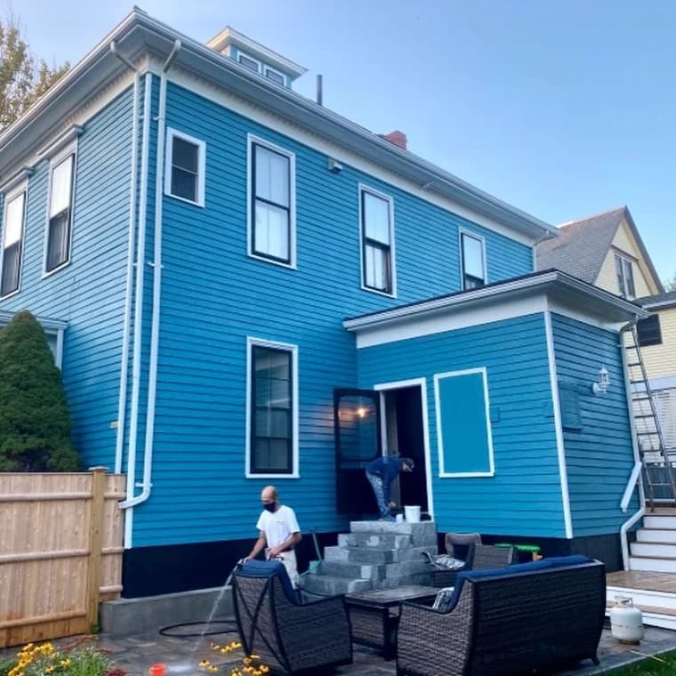 This is an image from Oliveira Painting Inc., located in Massachusetts. The photo shows a freshly painted two-story house with bright blue siding, white trim, and black accents, with workers completing the exterior project.