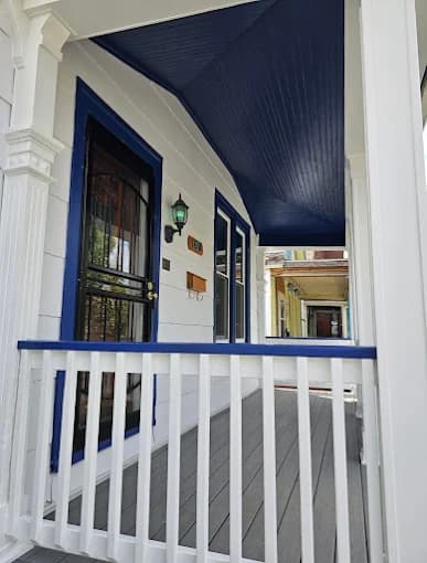 This is an image from BEE Pro Painting, located in New Jersey. The image shows a newly painted front porch with white railings, blue ceiling accents, and clean trim details surrounding the entryway.