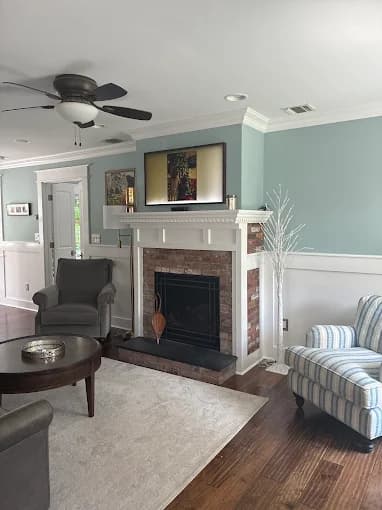This is an image from Elite Painting Services Plus, located in New York. The living room features light blue walls, white wainscoting, a brick fireplace with a white mantle, and coordinated seating in a bright, clean interior.