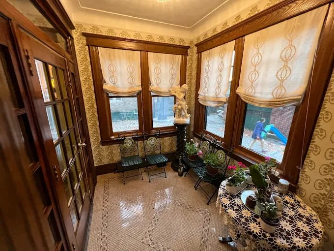 This is an image from By Hand Painting LLC, located in New Jersey. The photo shows a sunroom with patterned wallpaper, wood-trimmed windows, decorative curtains, indoor plants, and a small table with a lace cover.