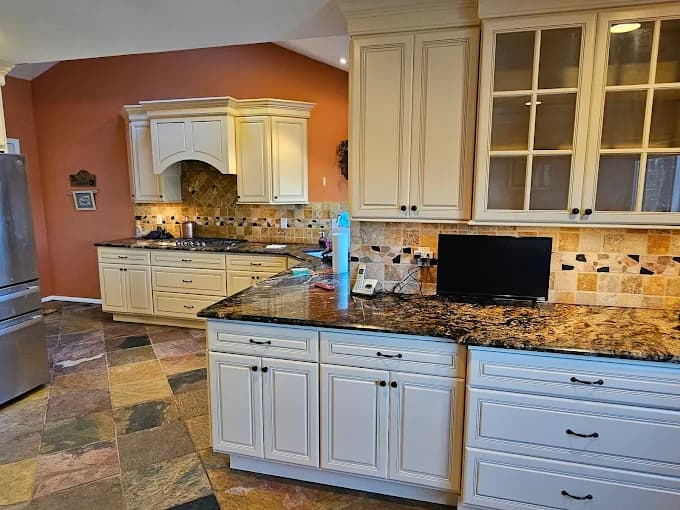 This is an image from Willow Tree Painting & Home Improvement LLC, located in New York. The kitchen features cream-colored cabinets, dark stone countertops, a multicolored tile backsplash, and a warm terracotta accent wall.