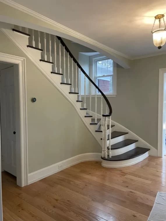 This is an image from Azul Painting Corp, located in New Jersey. The image shows a freshly painted residential interior featuring a curved staircase with white balusters, dark wood treads, and light neutral wall colors.