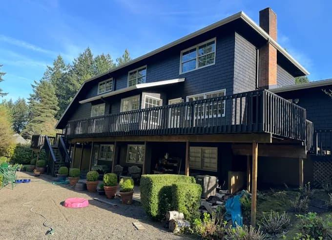 This is an image from Muir Painting Inc  company, located inHappy Valley, OR The image is a large two-story home with dark gray exterior paint, black deck railing, and multiple windows, freshly painted by Muir Painting Inc.