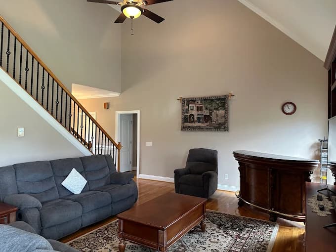 Interior painting project by Roca's Painters LLC in Mebane, North Carolina. The image shows a living room with gray sofas, a vaulted ceiling, and stair railing.