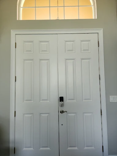 This is an image from Paint Masters Painting Contractors LLC company, located in Venice, FL. The image is a freshly painted white double entry doors with modern hardware and transom window in a light gray interior by Paint Masters Painting Contractors LLC