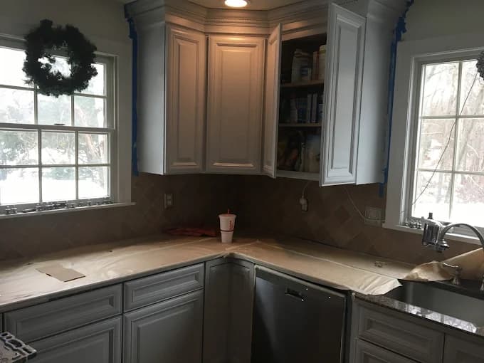 This is an image from CertaPro Painters of Woburn, Massachusetts. The photo shows a kitchen with freshly painted white cabinets, a tiled backsplash, and countertops prepared for refinishing, illustrating detailed interior painting work.