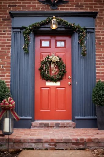 This is an image from Kennedy Painting Interior & Exterior, located in Indiana. The image shows a painted front entry door with a bold red finish, dark blue trim, brick surroundings, and decorative holiday accents highlighting detailed exterior painting work.