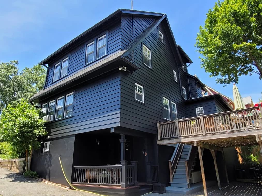 This is an image from Mark Tirondola Jr. Painting Contractor LLC in New Jersey. It shows a large three-story black house with white window trim and a wooden deck.