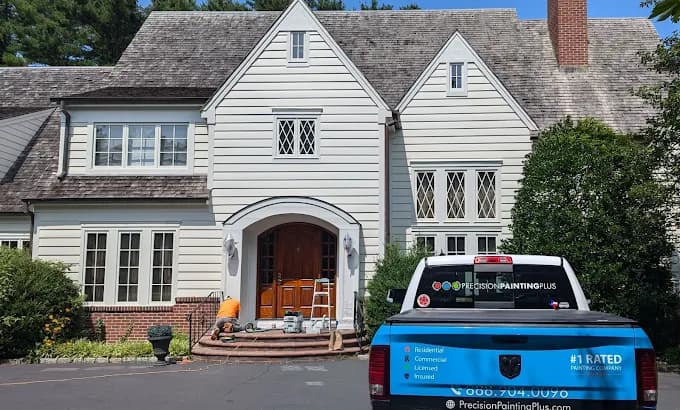 This is an image from Precision Painting Plus of Suffolk County, located in New York. The photo shows a professional painting crew working on a large white two-story home with a company-branded pickup truck parked in the driveway.