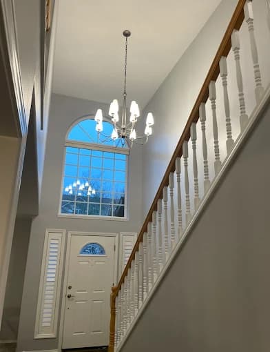 This is an image from Muir Painting Inc  company, located inHappy Valley, OR The image is an interior staircase and entryway freshly painted in light gray with white trim and railing, modern chandelier, and large arched window – Muir Painting Inc professional interior painting services.