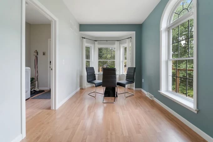This is an image from Brushes Over Broome LLC, located in New York. The interior photo shows a bright dining nook with freshly painted teal and light gray walls, large windows, and a small round table with modern black chairs.