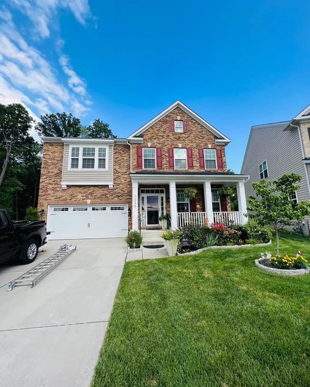 This is an image from Santana Painting Co, located in Maryland. The exterior view features a brick two-story home with red shutters, white trim, a landscaped front yard, and a double garage.