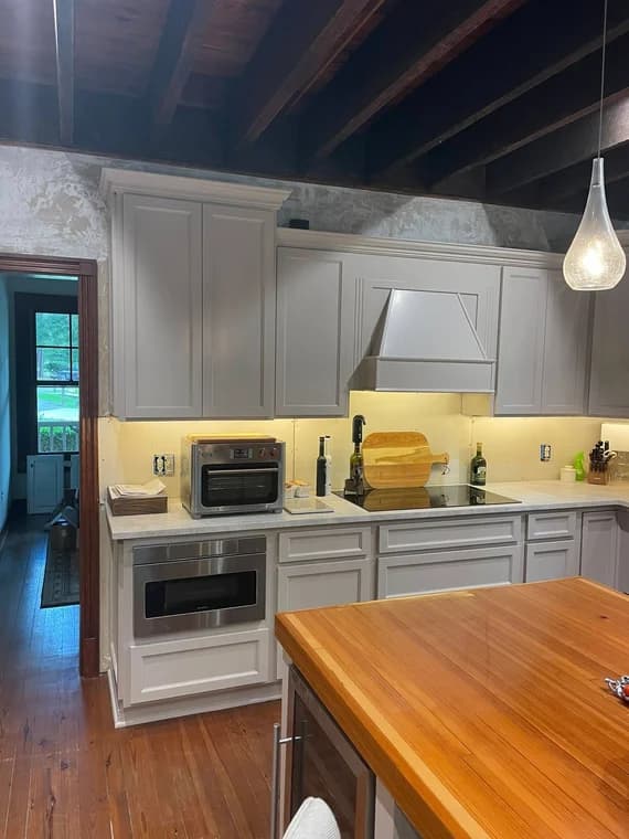 This is an image from Ponderosa Painting LLC, located in  Louisiana. The photo shows a modern kitchen interior with freshly painted light gray cabinets, a wooden island countertop, and exposed dark ceiling beams, highlighting detailed interior painting work.