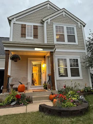 Exterior painting project by Metanoia Construction Inc. in Elburn, Illinois. The image shows a two-story home with neutral siding, white trim, wood shutters, and planters.