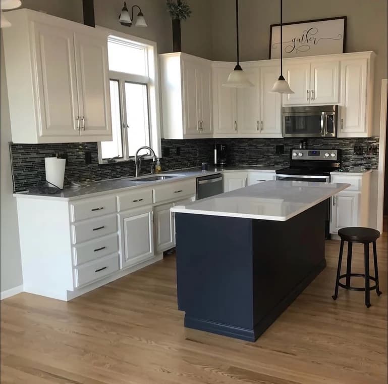 This is an image from Santana Painting Co, located in Maryland. The interior kitchen image shows white upper and lower cabinets, a dark navy island, a tiled backsplash, and natural hardwood flooring.