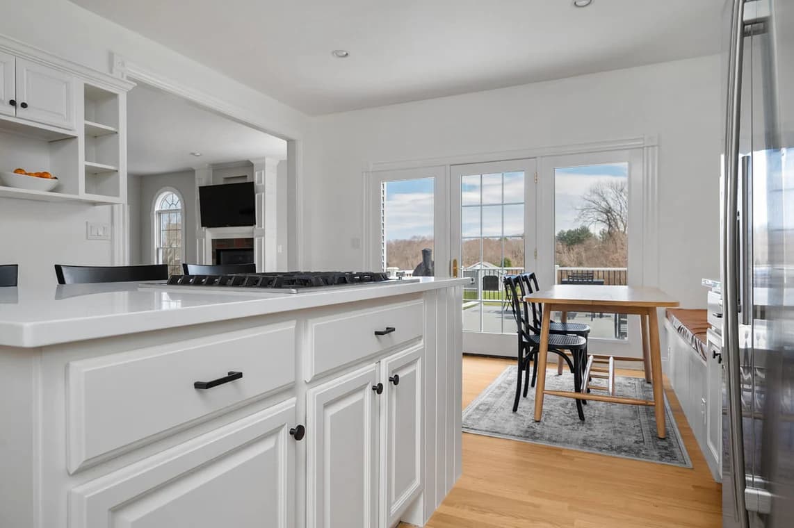 This is an image from AH Painting Services, located in Massachusetts. The photo shows a bright, freshly painted kitchen with white cabinetry, modern appliances, and large glass doors opening to an outdoor view, reflecting the company’s skill in creating clean and elegant interior finishes.