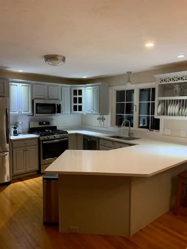 This is an image from DiNapoli Painting, located in Massachusetts. The photo shows a freshly painted kitchen with light gray cabinets, stainless steel appliances, white countertops, and wood flooring.