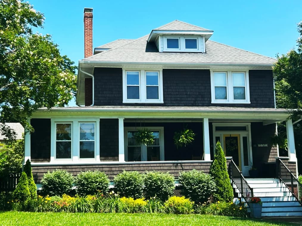 This is an image from Hands On Painters, located in Maryland. The exterior photo shows a large dark-shingled home with white trim, a full front porch, manicured landscaping, and a bright blue sky.