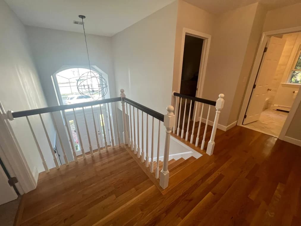 This is an image from Powell Painting & Home Services, located in Massachusetts. The photo shows a freshly painted upstairs hallway with neutral-toned walls, hardwood floors, white trim, and a modern chandelier above the entryway.