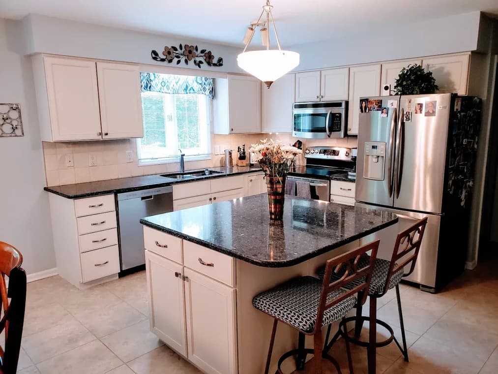 This is an image from Shabby Brown Restore, located in New York. The image displays a bright kitchen with white cabinets, stainless-steel appliances, and a large granite island centerpiece.