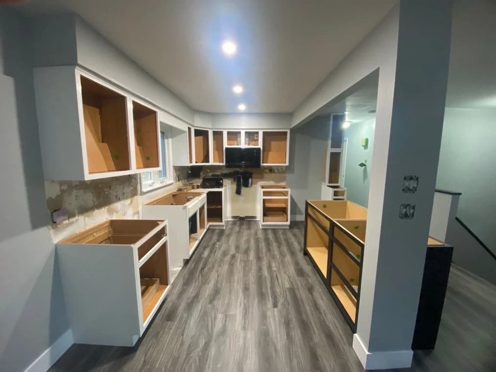 This is an image from Genesis Pro Painting & Restoration Inc, located in New York. The image shows a kitchen under renovation with cabinet frames installed, gray flooring, and recessed lighting above.