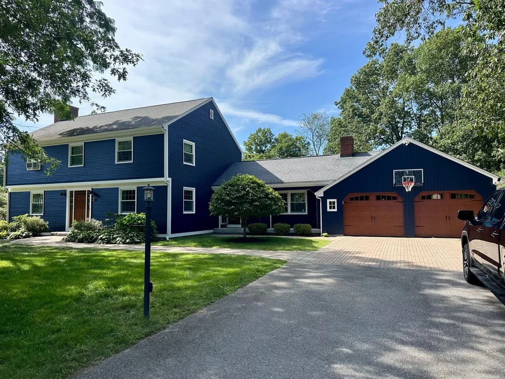 A two-story navy blue home with white trim and wooden garage doors, set on a spacious lot with a driveway and trees in the background.