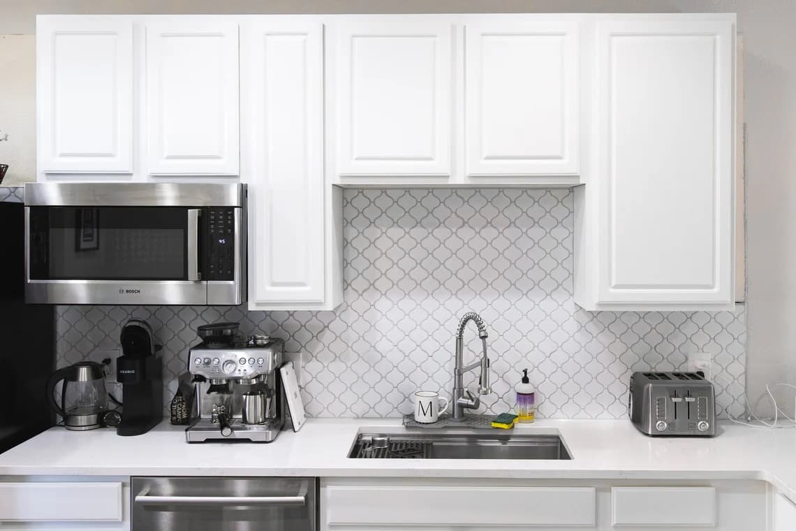 This is an image from Paint Denver company, located in Denver, CO The image is a modern kitchen with freshly painted white cabinets, stainless steel appliances, and patterned tile backsplash by Paint Denver