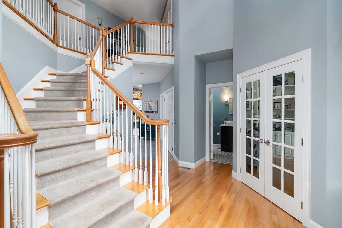 This is an image from AH Painting Services, located in Massachusetts. The photo shows a freshly painted interior entryway with soft blue-gray walls, white trim, and a staircase featuring light carpet and natural wood railings, highlighting the company’s detailed interior painting work.