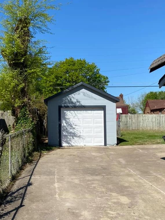 This is an image from Majestic Home Painting, showing a small gray garage with a white overhead door. The driveway is wide and open, bordered by a wooden fence and green grass.