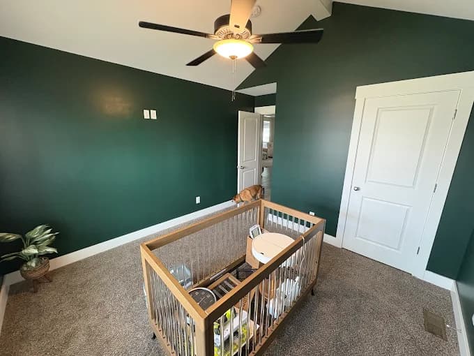 This is an image from R.E.A.L. Painting, located in Kentucky. The photo shows a nursery room with dark green walls, white trim, a ceiling fan, and a wooden crib.