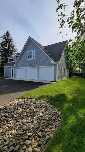 This is an image from Seven Oaks Painting Services, located in Ohio. The image displays a gray shingle-style home with white garage doors and a landscaped yard under bright daylight.