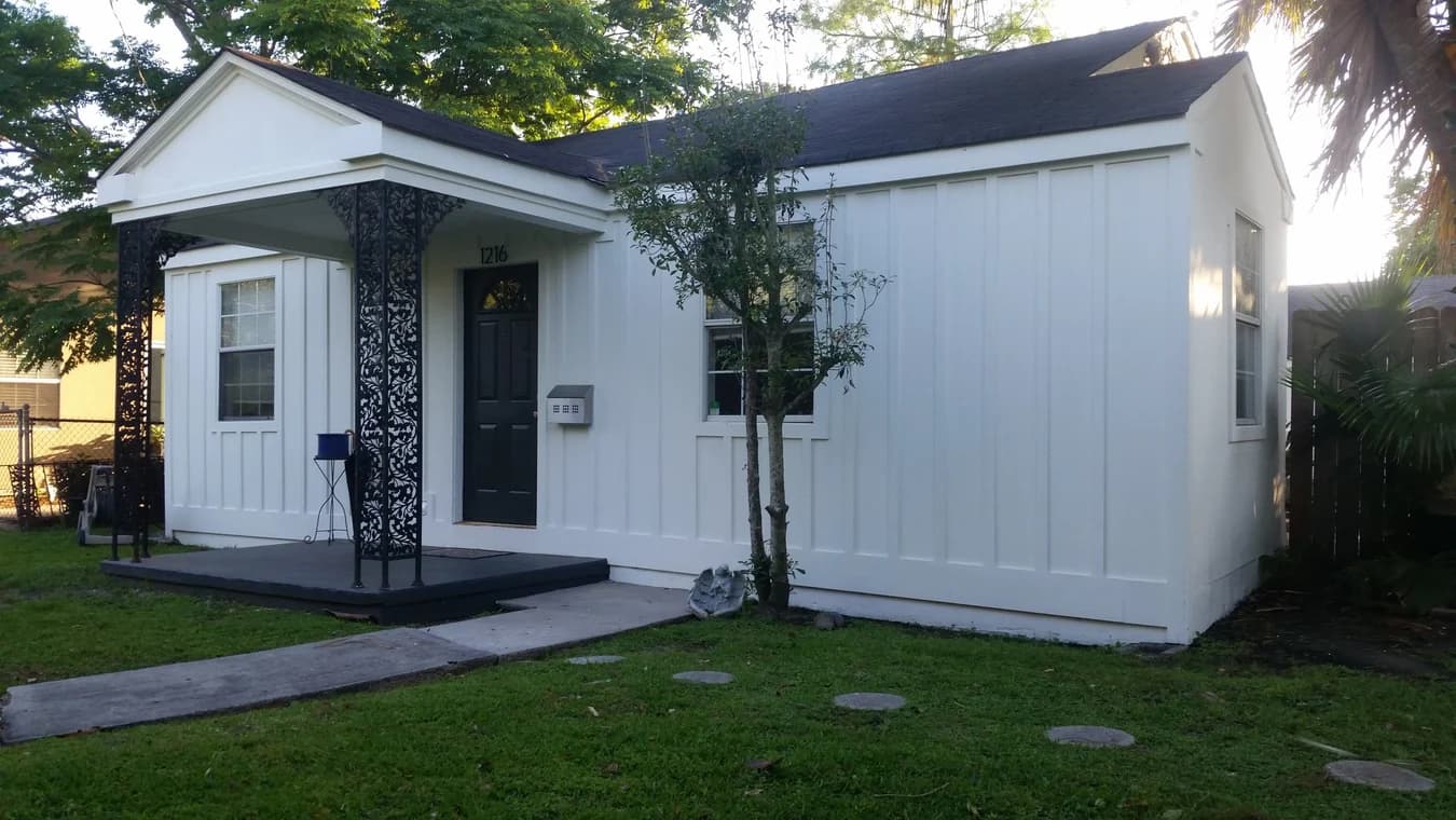 This is an image from Southern Dropcloth Painting LLC, located in Louisiana. The image shows a small white house with board-and-batten siding, a dark front door, and decorative black wrought-iron porch columns, surrounded by a green lawn and trees.