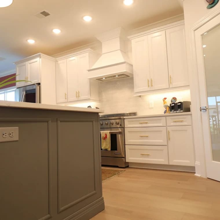 This is an image from Klappenberger & Son, located in Maryland. The photo shows a renovated kitchen with white cabinets, brass hardware, a custom range hood, and a gray island.