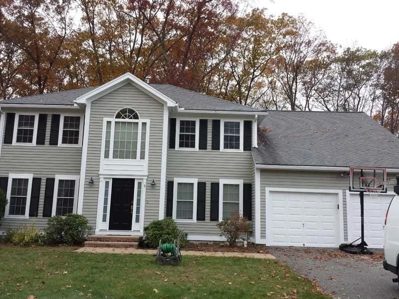 This is an image from Decolores Painting, showing a two-story gray house with black shutters and a black front door. The home has a large white garage and sits in front of a backdrop of autumn trees.