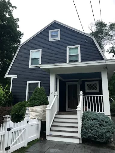 This is an image from DiNapoli Painting, located in Massachusetts. The photo shows a dark blue two-story home with crisp white trim, a front porch, and a white picket fence.