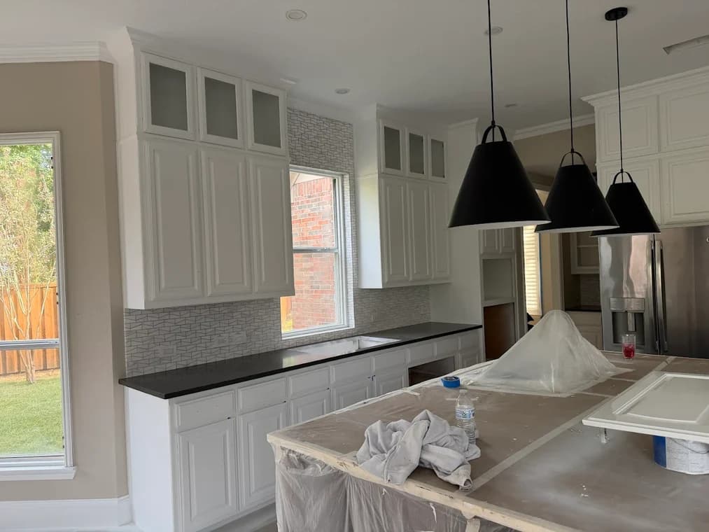 "This is an image from Hutch-N-Son Painting & Drywall, located in Texas. The photo shows a kitchen with freshly painted white cabinets, black countertops, and pendant lighting over a covered island during renovation."