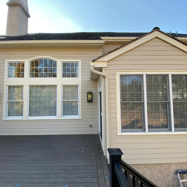 This is an image from Benner & Sons Inc company, located in Newtown Square, PA. The image is an exterior house painting project featuring tan siding with white trim, large grid-style windows, and composite deck with black railing.